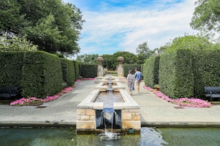 A man and woman walk beside a long, rectangular pond between well-kept bushes and small pink flowers at Dallas Arboretum