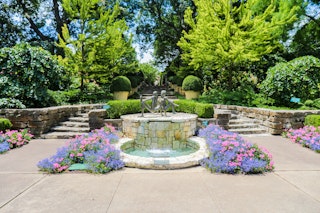 A statue and fountain surrounded by purple and pink flowers with two stone staircases on either side